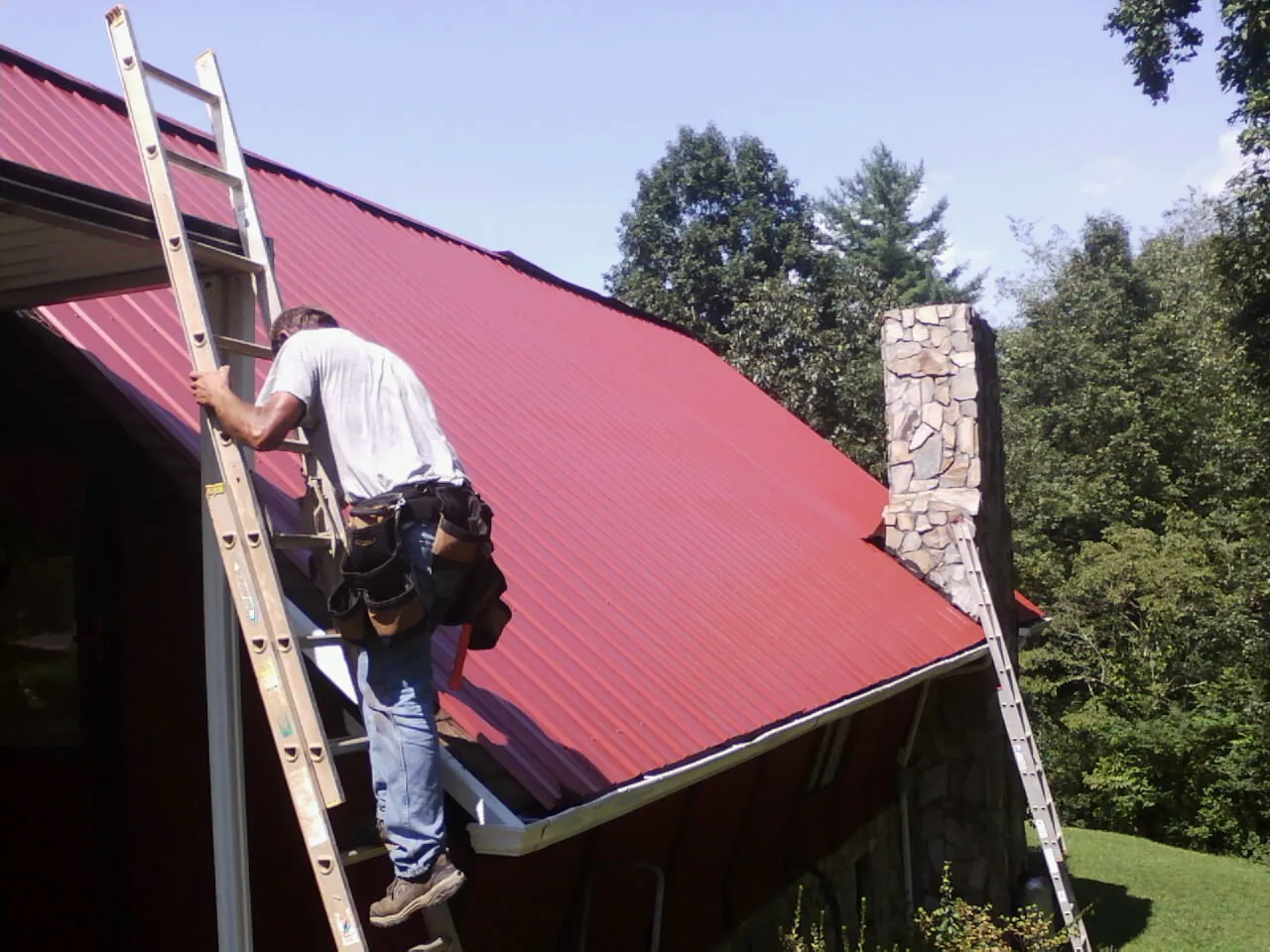 Roofer on a ladder beside a red metal roof and stone chimney — Soffit & Fascia Repair by Roofing Brothers in Fountainville